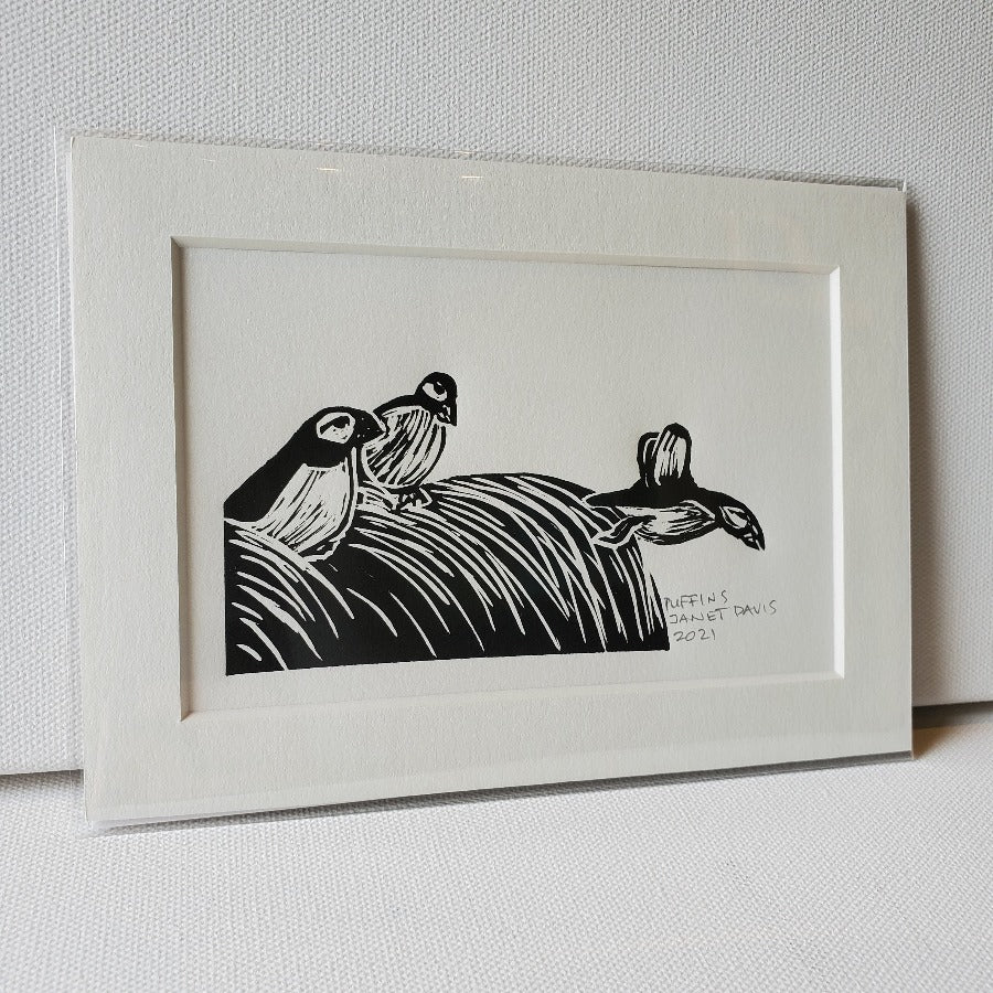 Three puffins: two seated, one flying off the edge of a grassy embankment. Lino-cut relief Mini-Print in black ink.