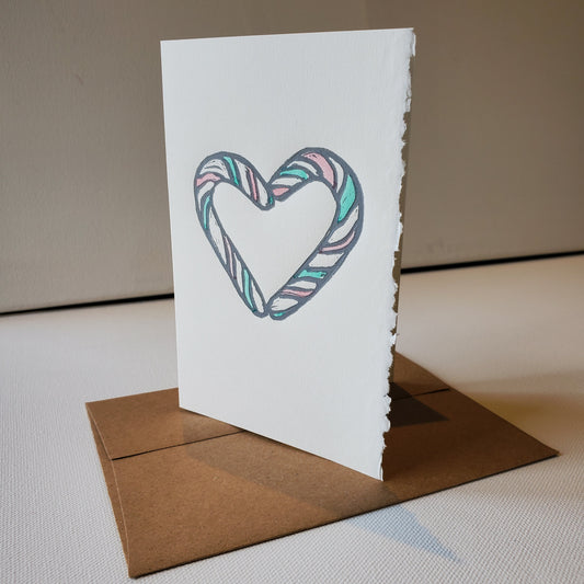 A 4 x 6 inch Christmas card with an candy cane heart design, made on antique white cotton fibre paper, with a natural decal edge. It features a hand-printed lino-cut and comes with a Kraft envelope.
