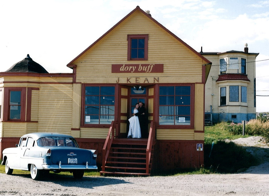 Janet & Duke on their wedding day, standing on the steps of the c.1890 Kean's General Store.  The shop is a wooden clapboard building with a peak roof, as well as a domed extension. The shop is painted dory buff. Photo credit to Edgar Blackwood.