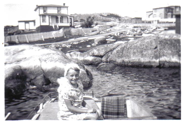 A young girl sitting in a boat in Norton's Cove with Captain Charlie Kean's house in the background.