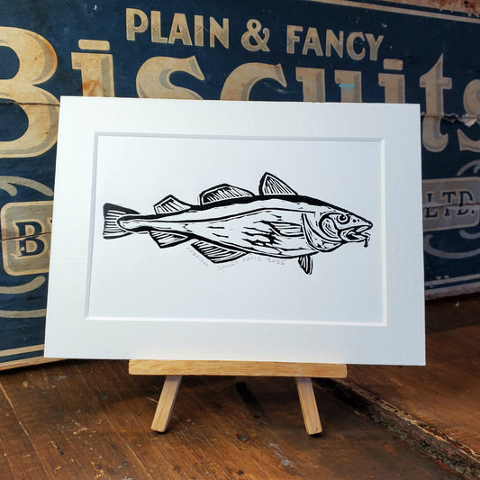A matted art print featuring a black ink illustration of a fish on a white background, displayed on a small wooden easel against a vintage-style biscuit box and wooden tabletop.