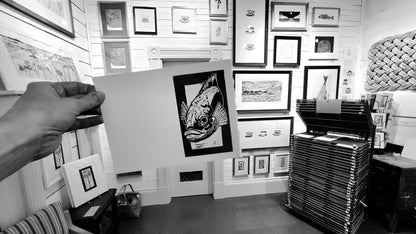 Person holding a freshly printed card at Norton's Cove Studio with various framed artworks on the walls, Black & White photo.
