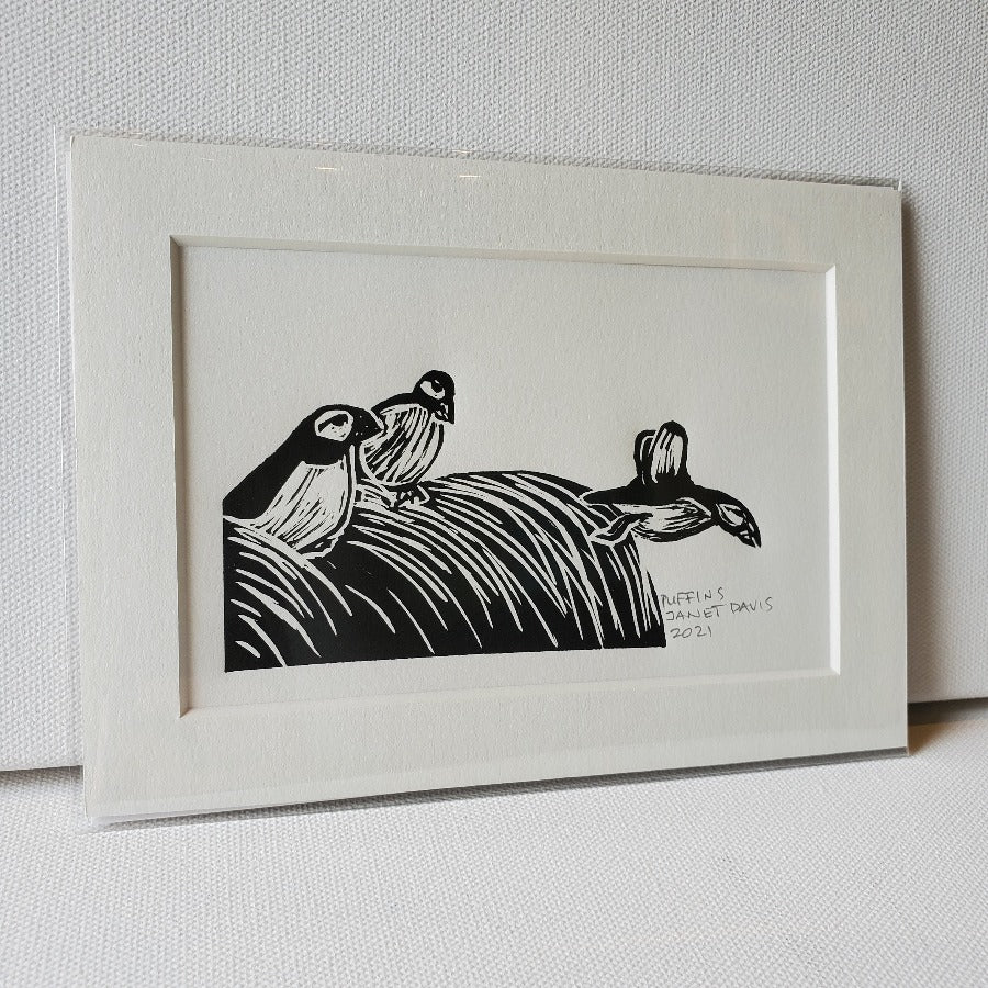 Three puffins: two seated, one flying off the edge of a grassy embankment.  Lino-cut relief Mini-Print in black ink.